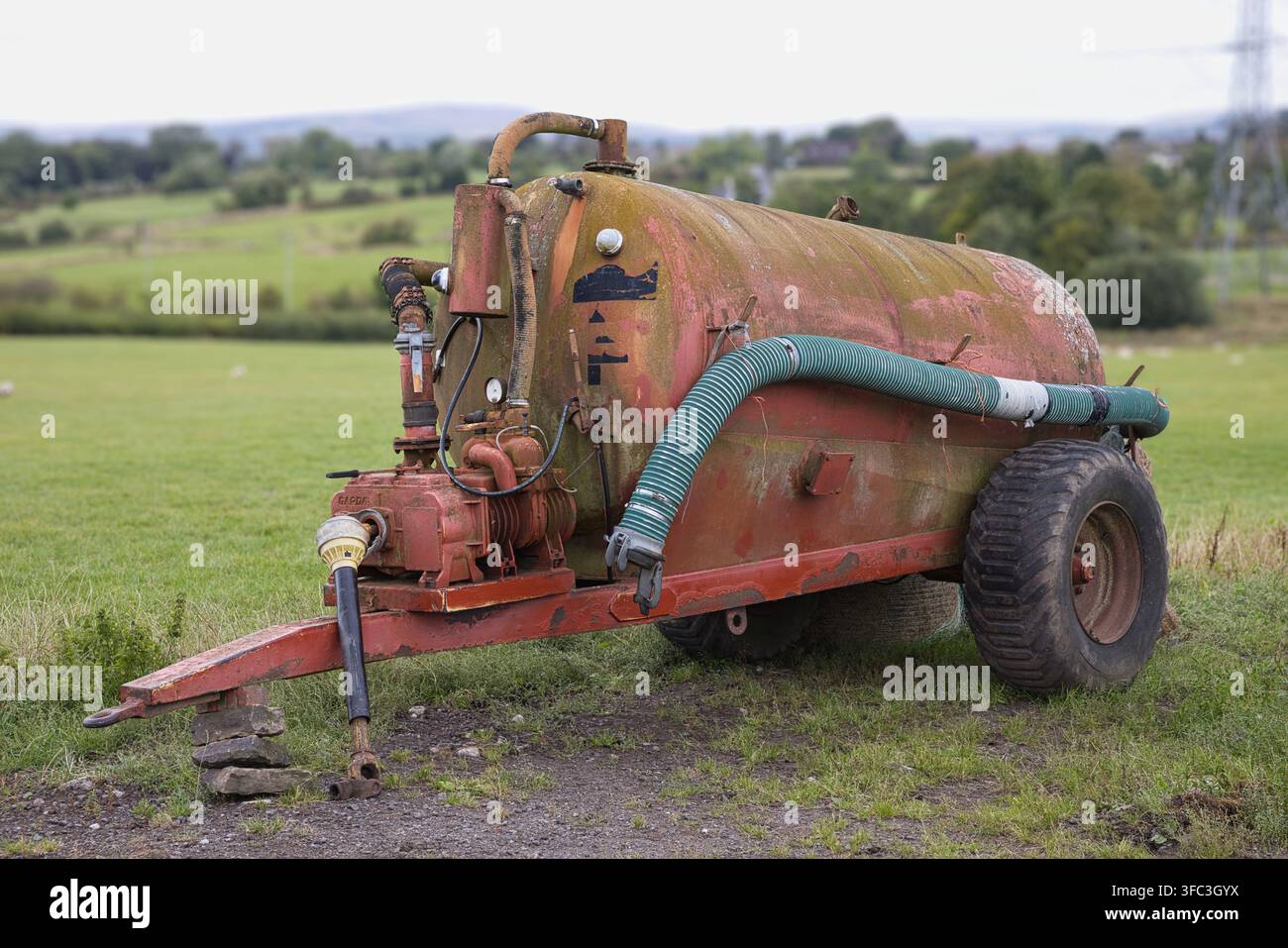 Remorque citerne à lisier rouge vintage garée dans un champ agricole vert, montrant une peinture érodée, de la rouille et un gros tuyau d'aspiration. Banque D'Images