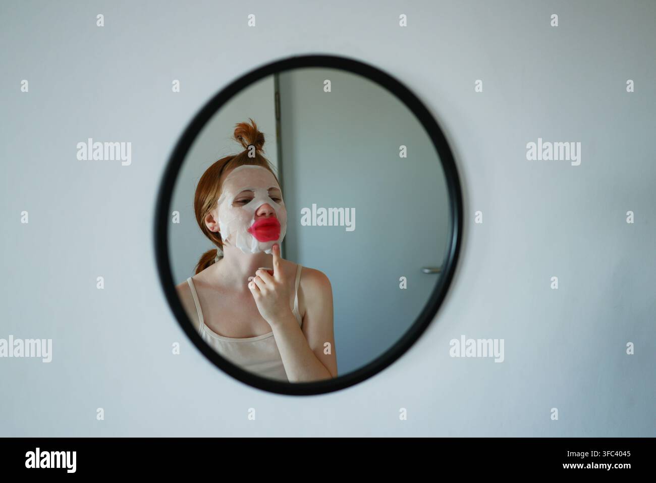 Routine de soins à domicile avec une jeune femme utilisant un masque hydratant pour le visage et un patch pour les lèvres tout en vérifiant son reflet dans un miroir rond. Banque D'Images
