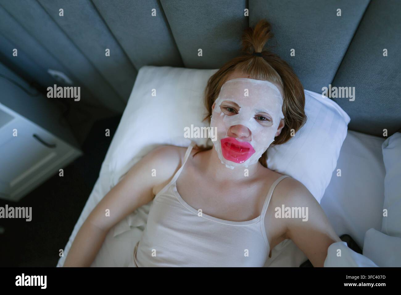 Femme allongée dans un lit portant un masque facial et un patch pour les lèvres, combinant soins de la peau, détente et mode de vie numérique. Banque D'Images