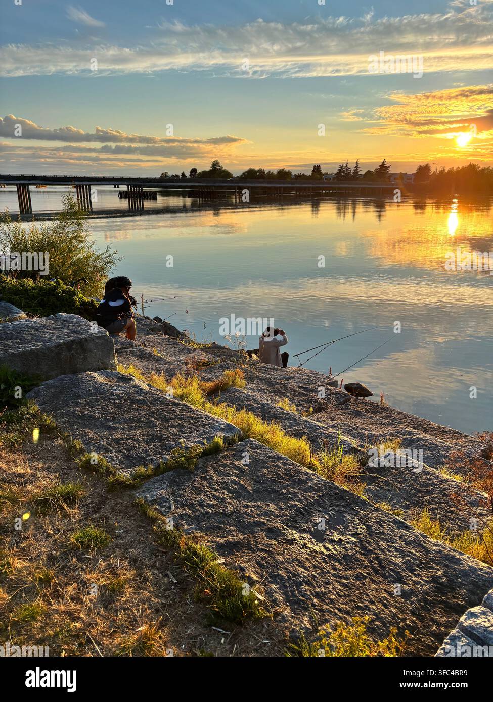 Deux personnes pêchent depuis une rive rocheuse au coucher du soleil, leurs cannes surplombant une eau calme baignée d'une lumière dorée - Image de stock capturée avec un smartphone