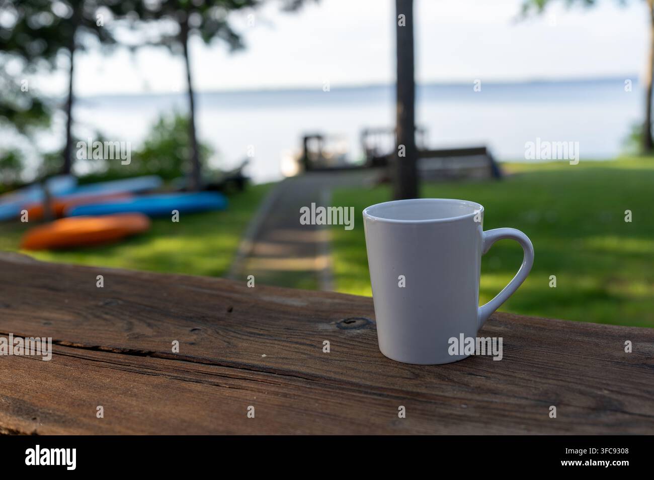 Close-up d'une tasse à café blanche sur une table en bois rustique surplombant un lac calme et une pelouse verte. Des kayaks colorés et une jetée sont doucement flous dans le b Banque D'Images