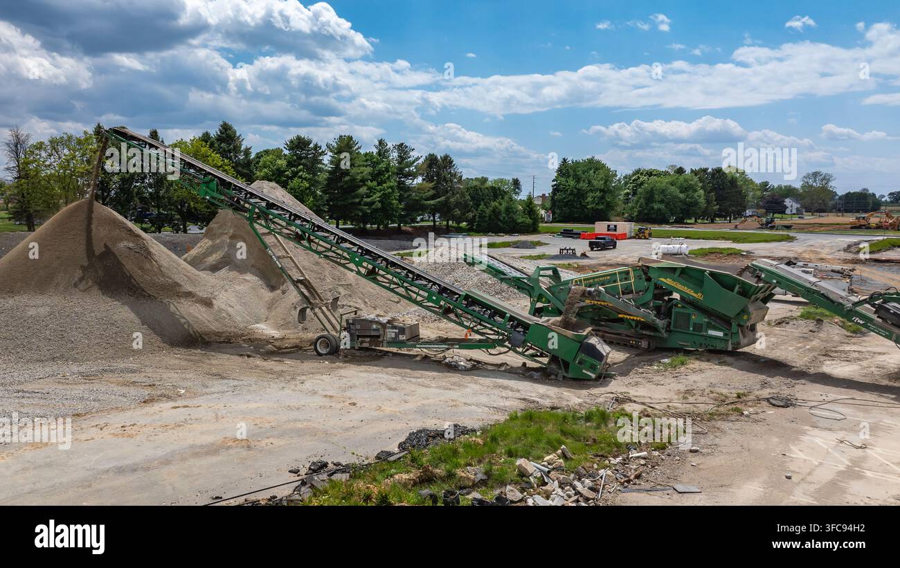 Des machines lourdes travaillent sur un chantier de construction pour traiter du gravier et créer des tas. L'équipement se fond dans le paysage naturel sous un ciel bleu avec Banque D'Images