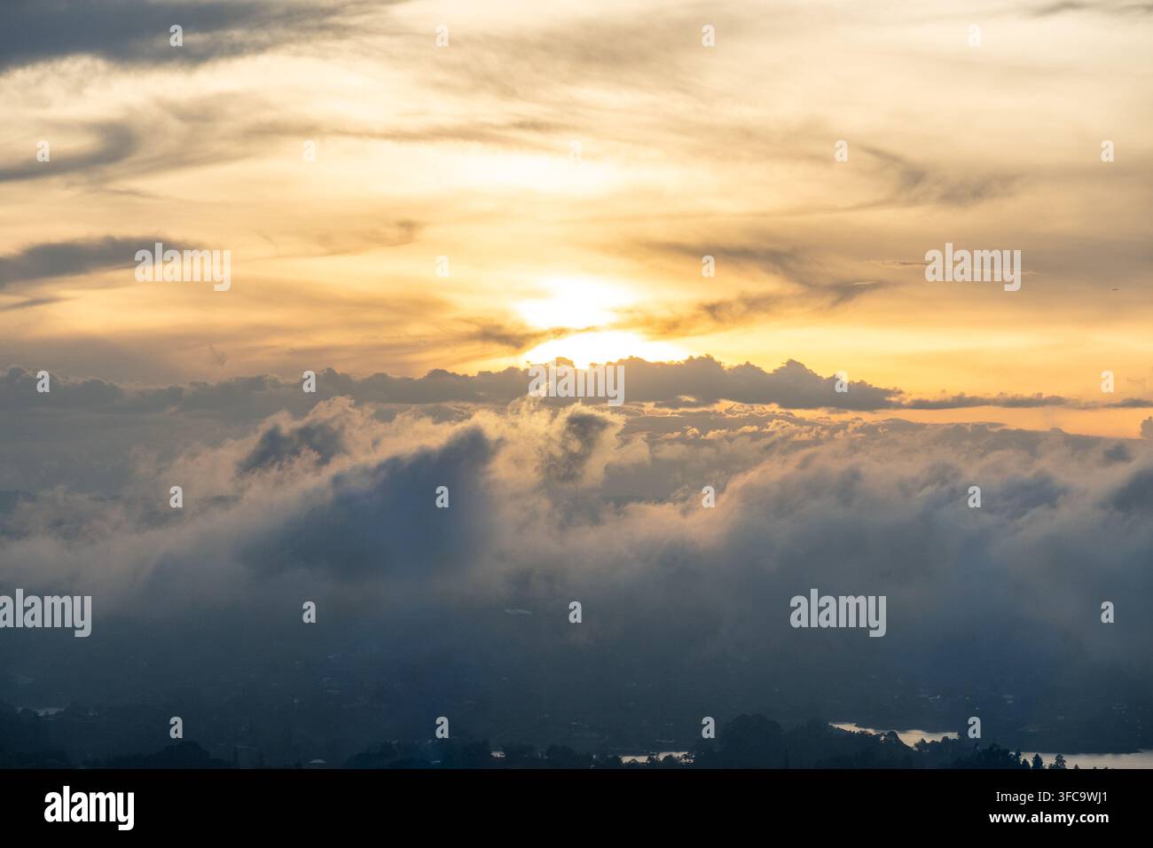 Lumière chaude dorée du soleil perçant un ciel spectaculaire de nuages superposés au-dessus de collines brumeuses et d'un lac faiblement visible. La scène montre une forma nuageuse à haute altitude Banque D'Images