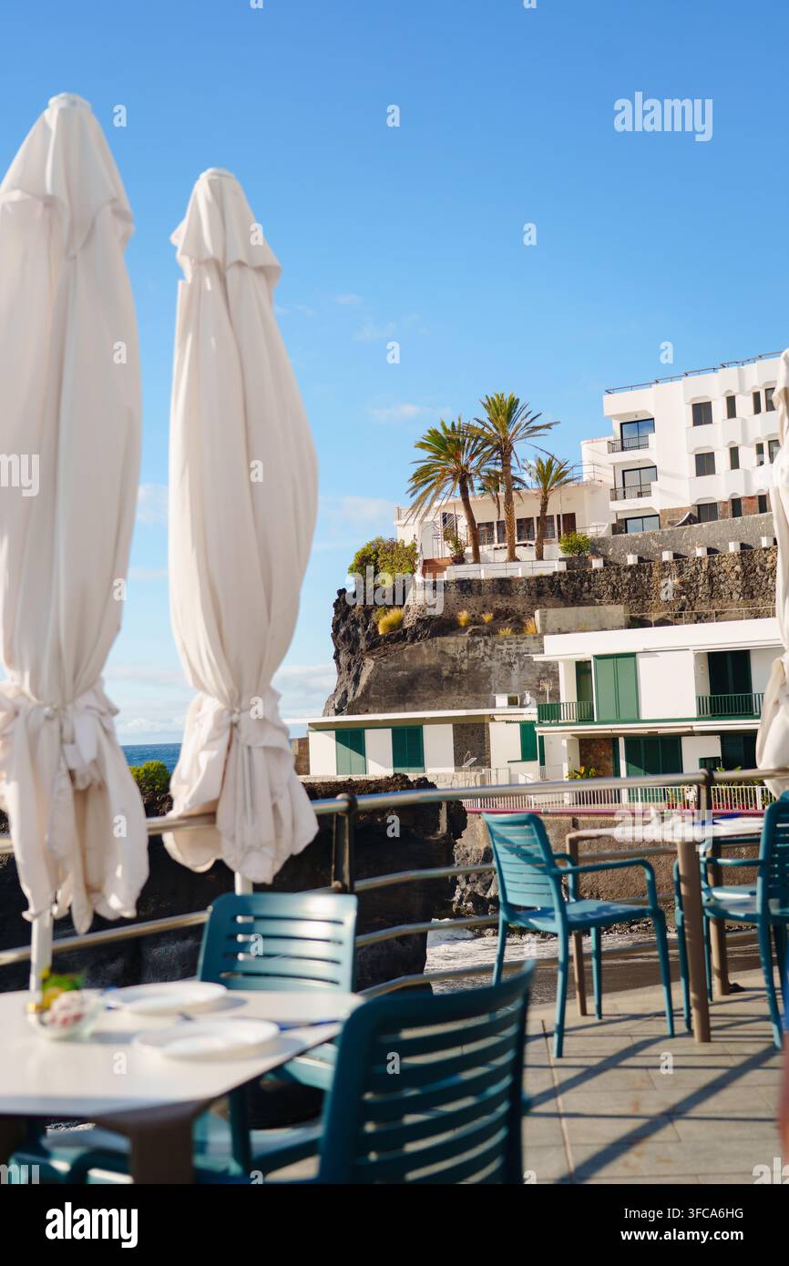 Parasols blancs repliés et chaises bleu-vert vides sur une terrasse de restaurant au sommet d'une falaise surplombant l'Atlantique, Puerto Naos, La Palma, Espagne Banque D'Images