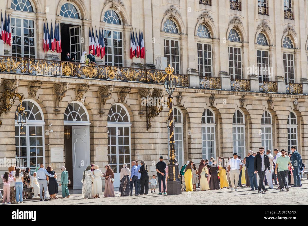 Des personnes en tenue festive se rassemblent devant la façade de style baroque avec ses rambardes dorées du balcon de l’Hôtel de Ville de Nancy sur la Rue Sainte-Catherine à Nancy, Grand Est, France. Un marié et une mariée sur le balcon saluent la foule en attente, tandis que plusieurs drapeaux tricolores pendent entre les fenêtres. Banque D'Images