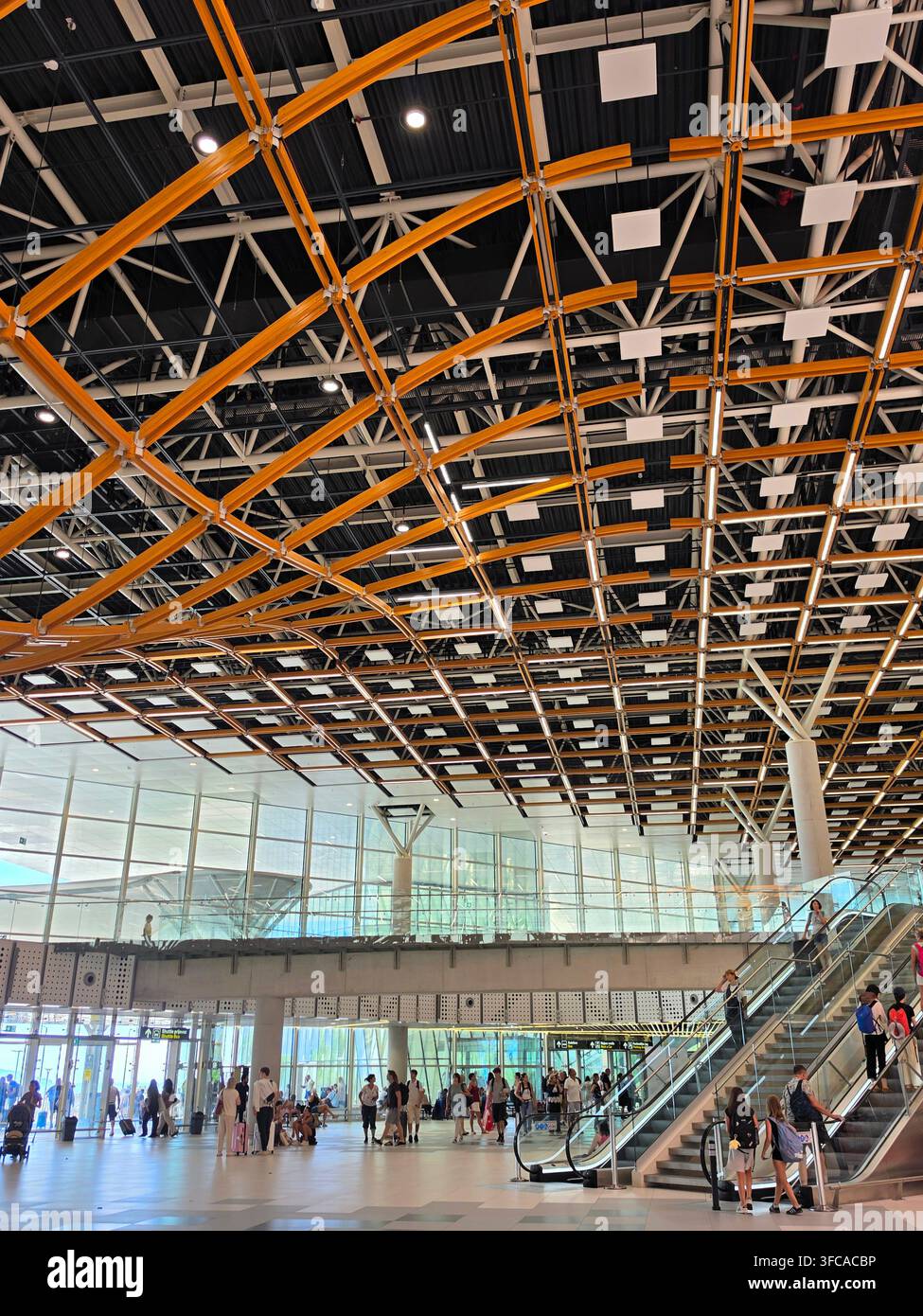 Intérieur moderne d'un terminal d'aéroport présentant une structure complexe de plafond orange et métallique, des escaliers mécaniques et des passagers marchant dans l'espace brillamment éclairé Banque D'Images