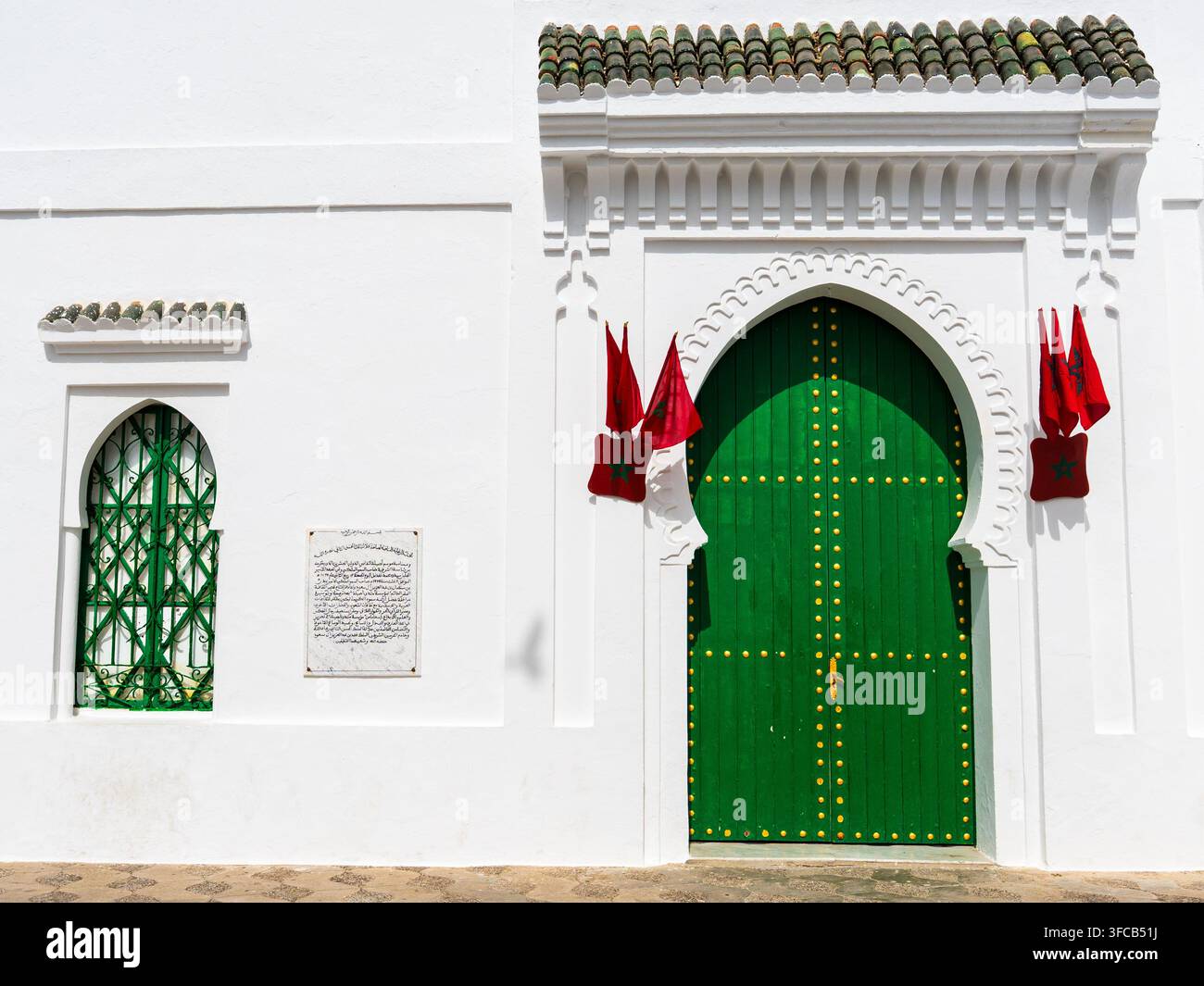 Façade de porte arquée en bois vert marocaine traditionnelle surmontée de tuiles céramiques vertes et flanquée de drapeaux marocains dans le medina historique d'Asilah, Maroc Banque D'Images