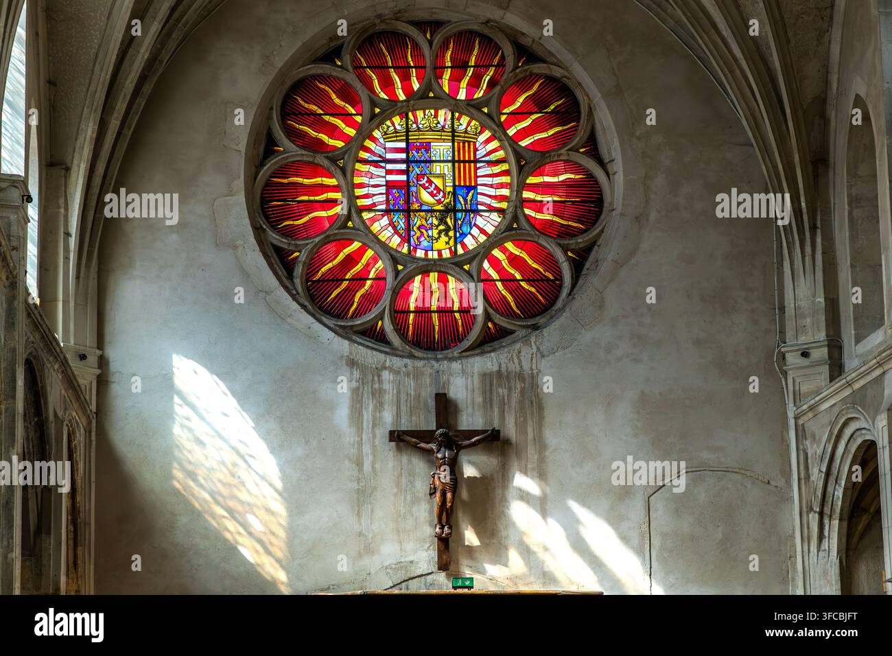 Un crucifix est suspendu sous le rosace portant les armoiries des ducs de Lorraine dans l'église des Cordeliers à Nancy, Grand Est, France. La lumière du soleil tombe en biais à travers les vitres latérales sur le mur. Banque D'Images