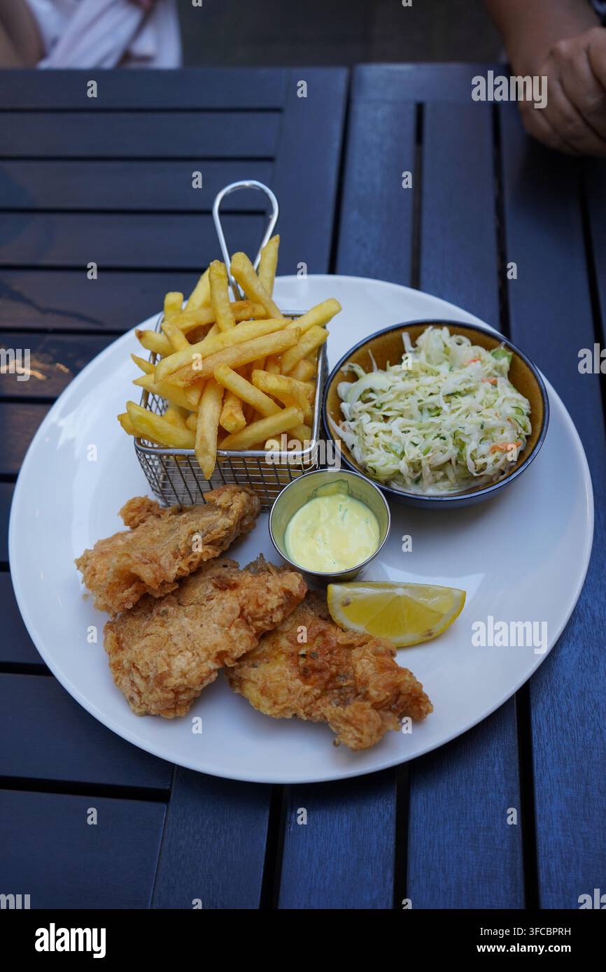 Prise verticale d'une assiette de filets de poisson frits panés croustillants servis avec des frites dorées dans un panier en métal, une salade de chou et une sauce tartare Banque D'Images