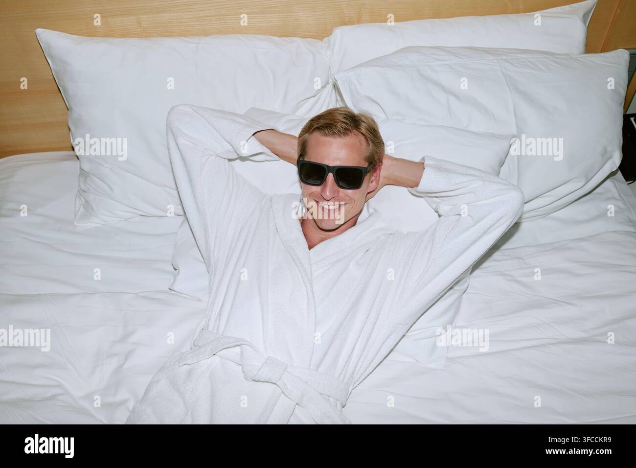 Portrait d'un homme souriant en peignoir et lunettes de soleil se détendant sur un lit d'hôtel Banque D'Images