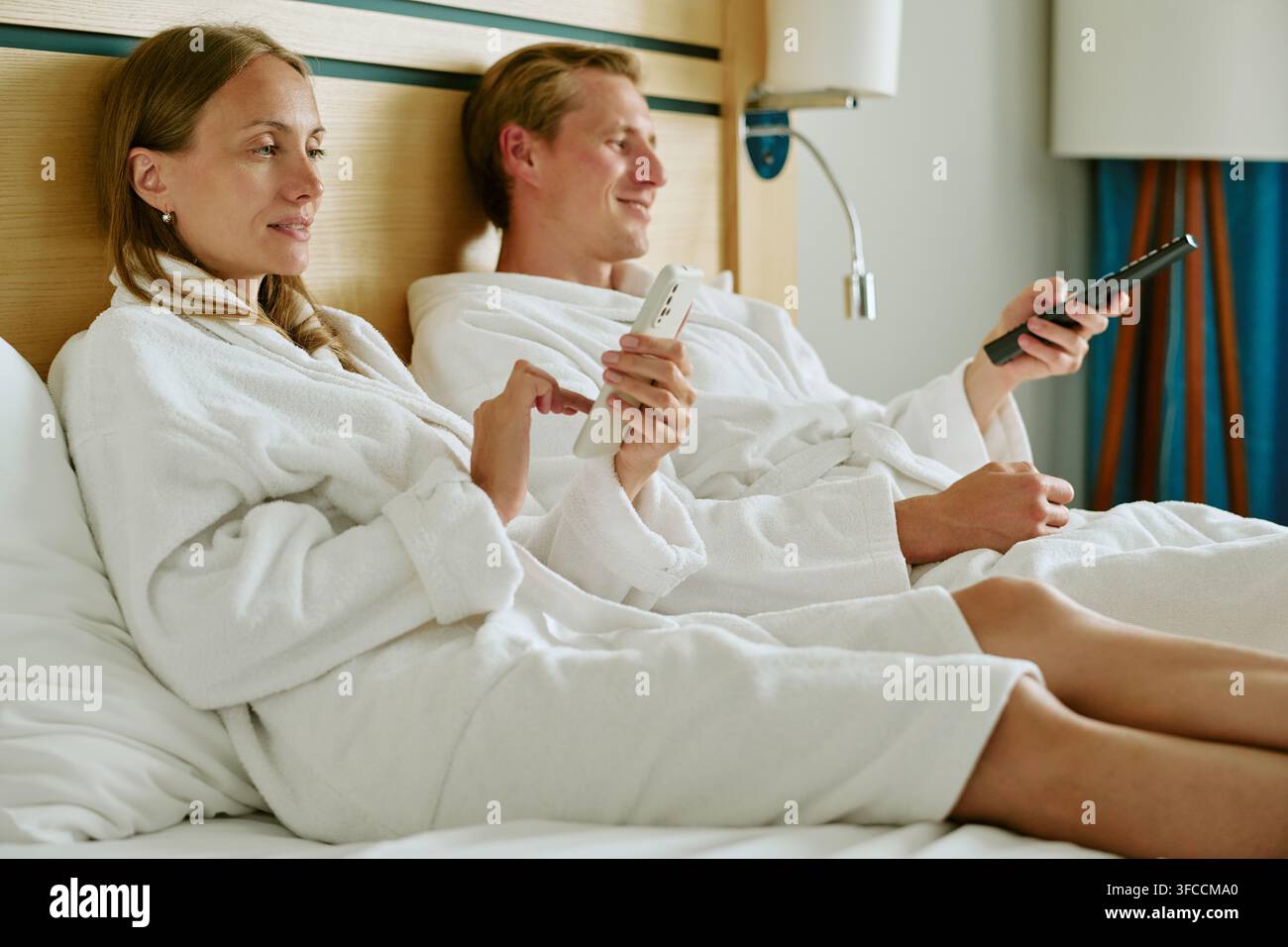 Couple en peignoirs se détendant dans un lit d'hôtel en utilisant un smartphone et une télécommande Banque D'Images