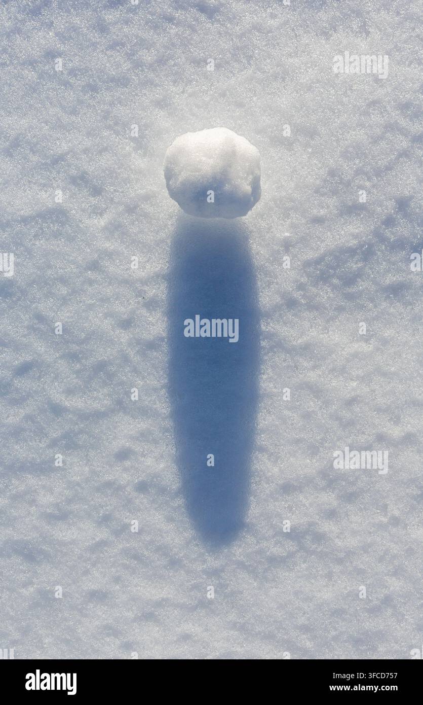 Une seule boule de neige blanche projetant une longue ombre bleue sur la neige texturée sous la lumière du coucher de soleil. Banque D'Images