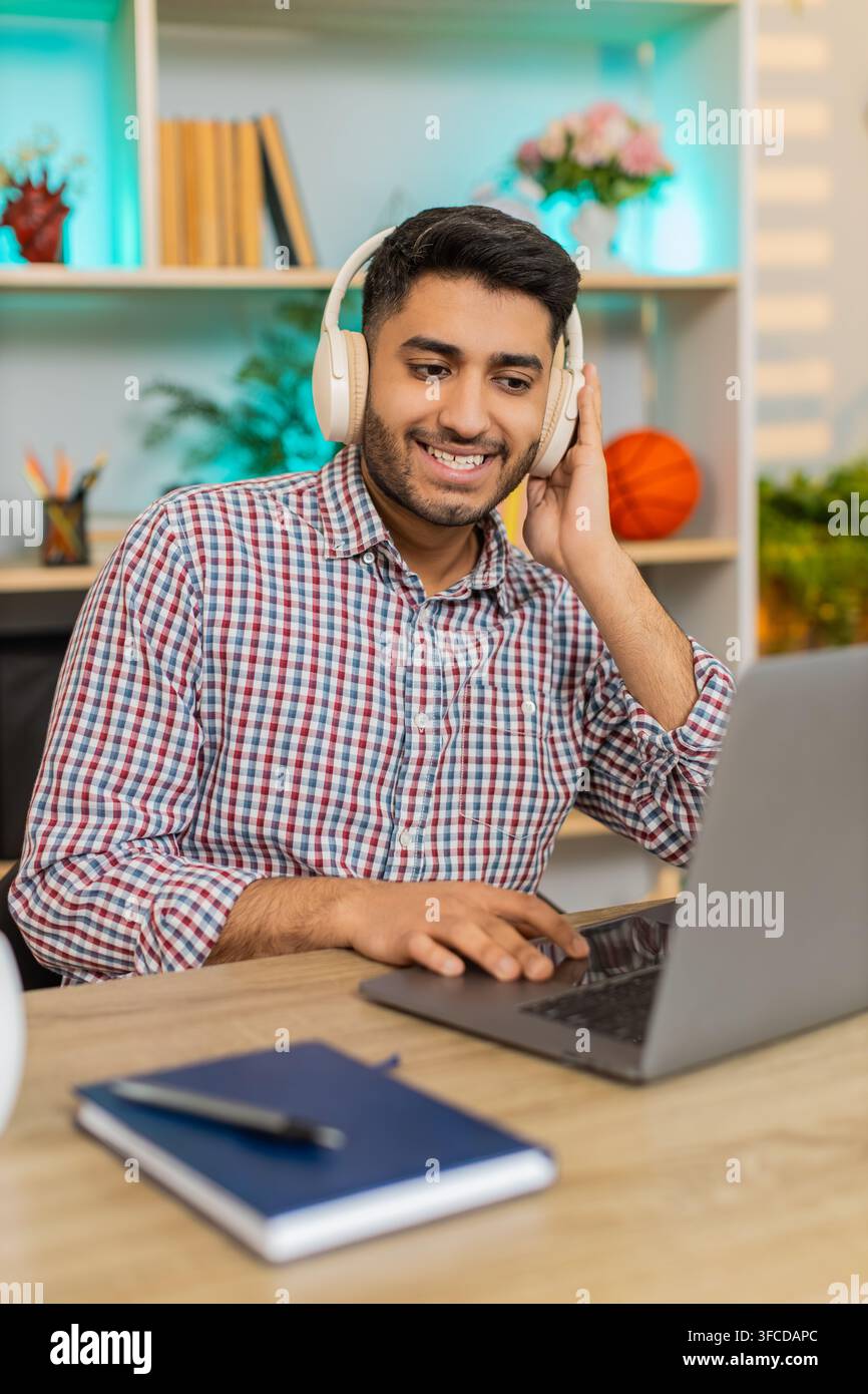 L'homme indien sur la table à la maison travaille sur ordinateur portable dans des écouteurs sans fil tapant du texte et écoutant de la musique avec une concentration calme à l'intérieur. Arabian Guy sur la table de la maison apprécie le travail freelance détendu et la routine de bureau à domicile Banque D'Images