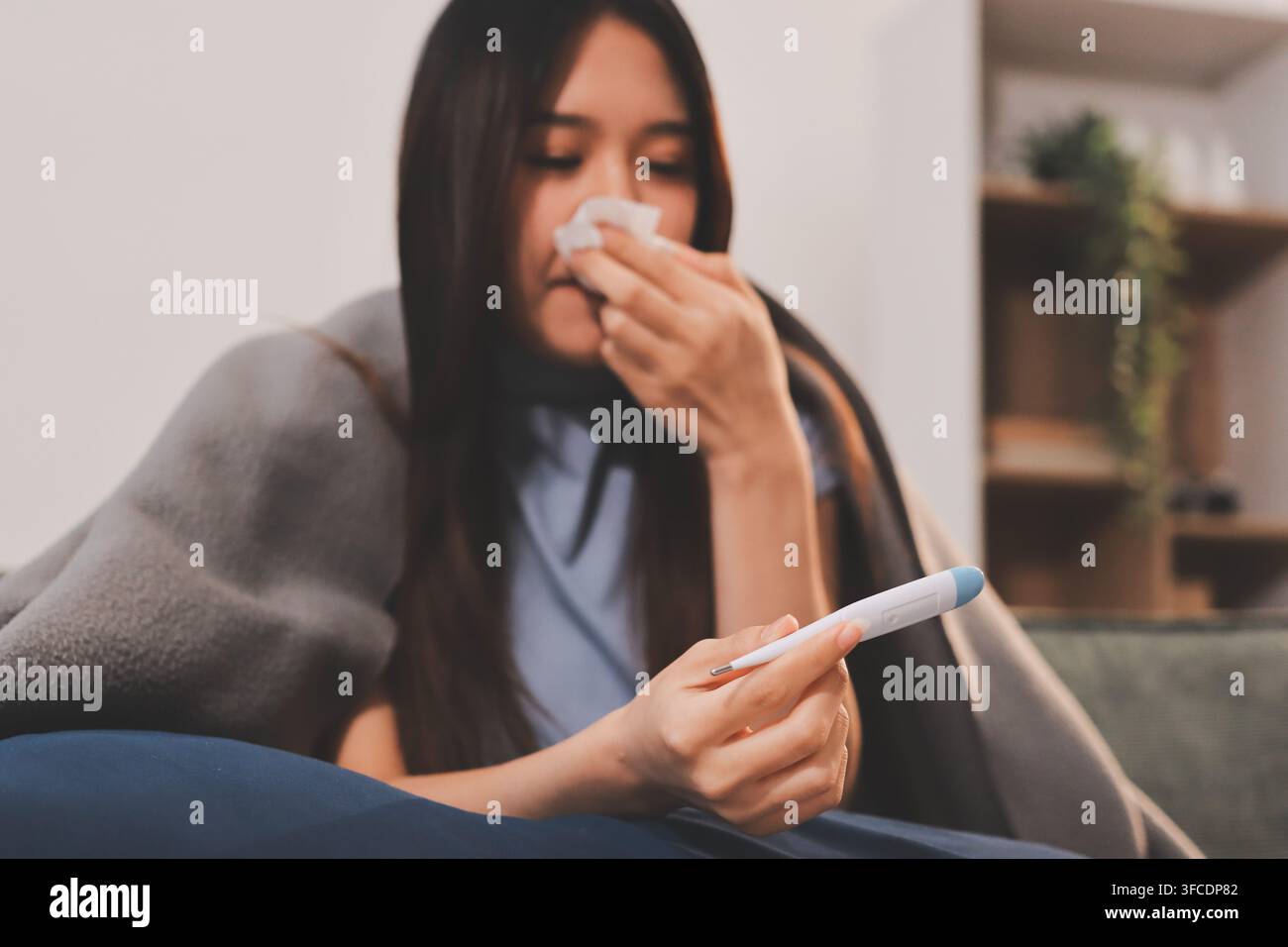 jeune femme asiatique malade éternuant dans un mouchoir en papier tout en étant recouverte d'une couverture et assise sur un canapé dans le salon Banque D'Images