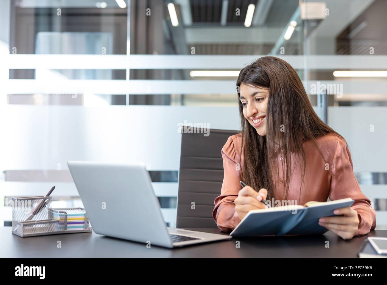 Jeune femme d'affaires souriante en train d'écrire dans un journal tout en utilisant un ordinateur portable, organisant des tâches et planifiant à un bureau d'entreprise moderne, concentrée, productive et confiante Banque D'Images