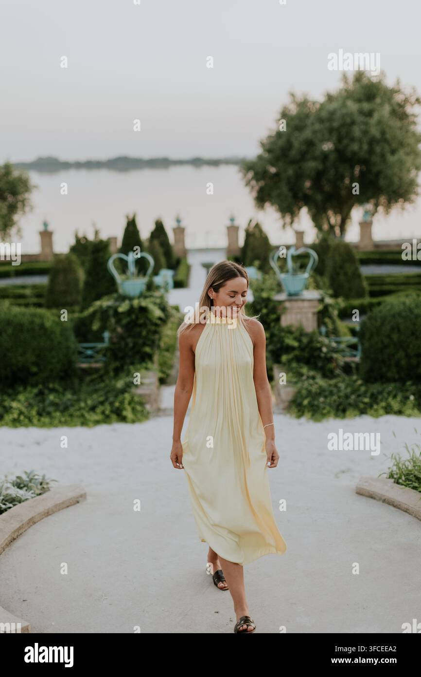 Une jeune femme dans une robe jaune fluide marche avec bonheur à travers un jardin luxuriant au coucher du soleil. Banque D'Images