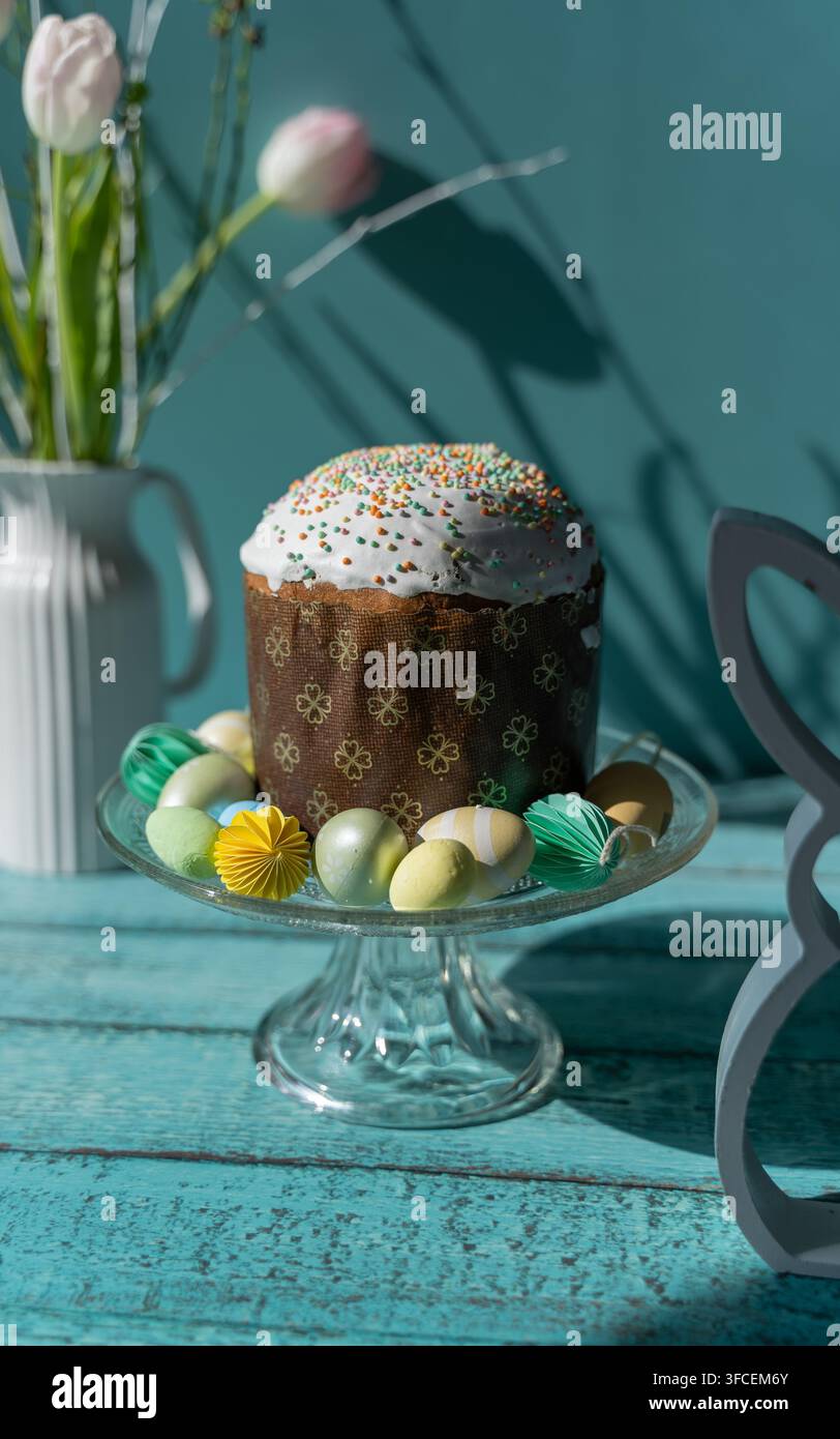 Pain de Pâques traditionnel Paska ou Pasca présenté sur un support à gâteau en verre avec des œufs de Pâques décoratifs et un ornement en forme de lapin sur un fond en bois bleu Banque D'Images
