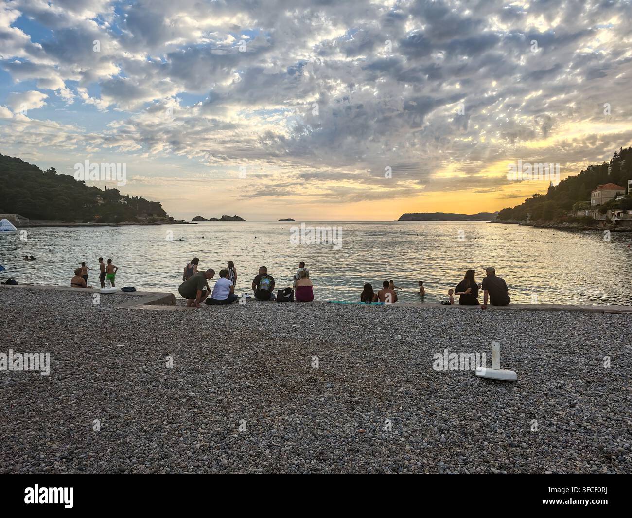 Des gens assis sur le rivage de galets de la plage de Lapad, profitant d'une vue sereine du coucher de soleil sur la mer Adriatique tranquille. - Image de stock capturée avec un smartphone