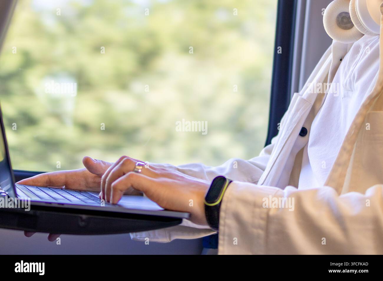 Professionnel utilisant un ordinateur portable à côté d'une fenêtre de train avec des écouteurs autour du cou. Concept de travail en mouvement, style de vie numérique, voyages d'affaires et mobilité Banque D'Images