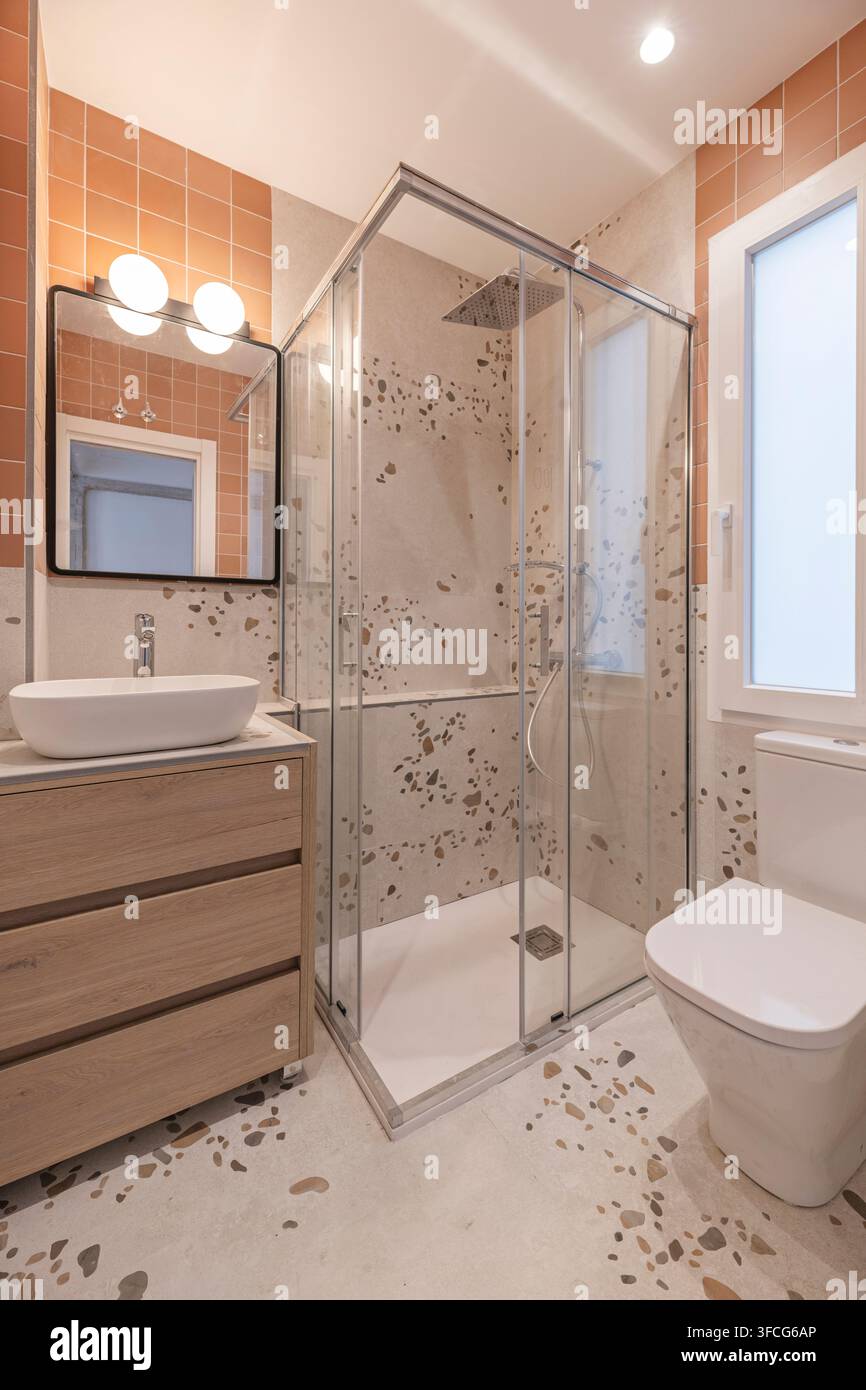 Salle de bain moderne carrelée en terrazzo avec douche d'angle en verre, pommeau de douche effet pluie, lavabo sur pied, meuble de toilette en bois et murs en terre cuite. Banque D'Images