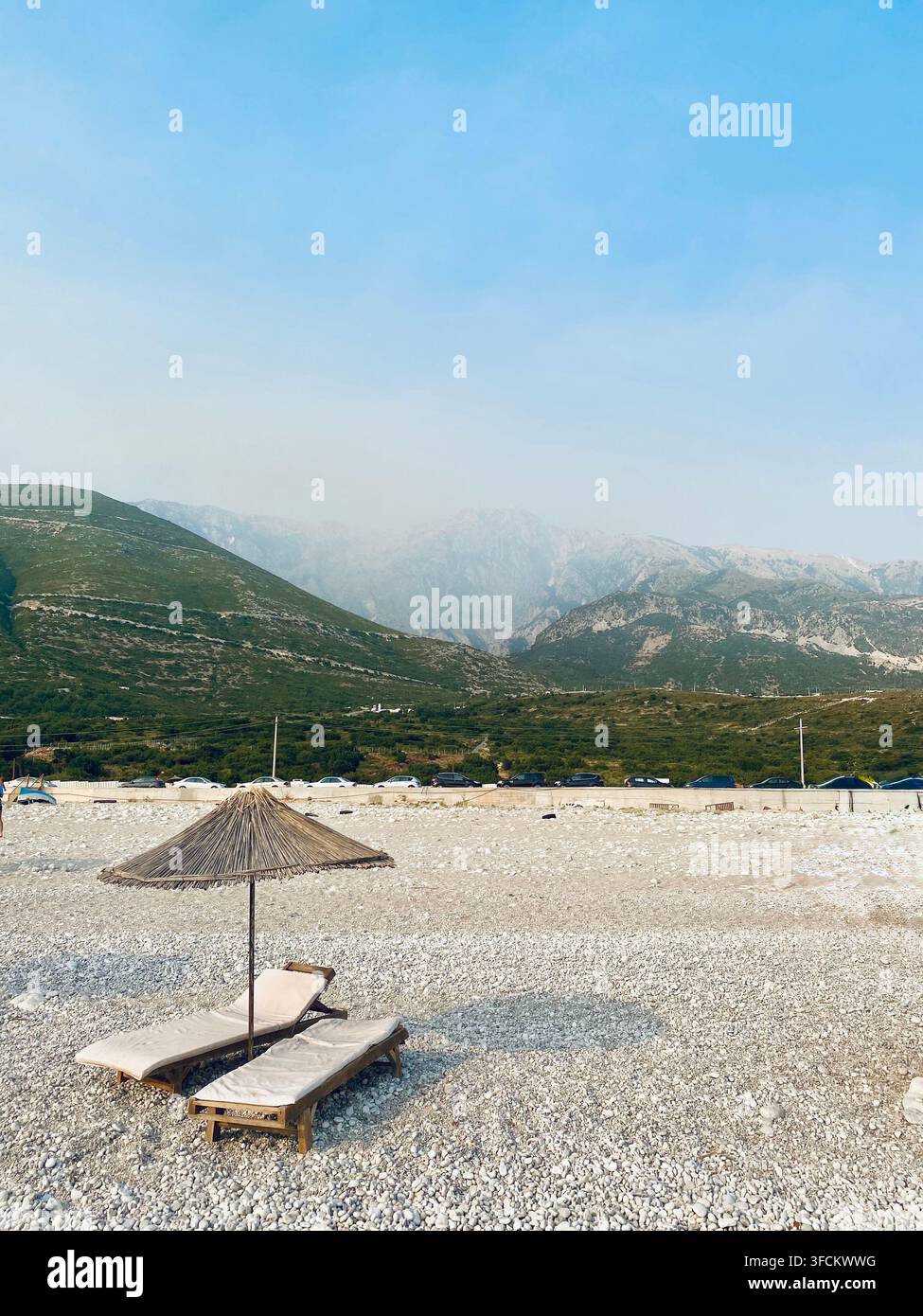 Deux chaises longues et un parasol en paille se trouvent sur une large plage de galets sous des collines vertes et un arrière-plan montagneux spectaculaire. Banque D'Images