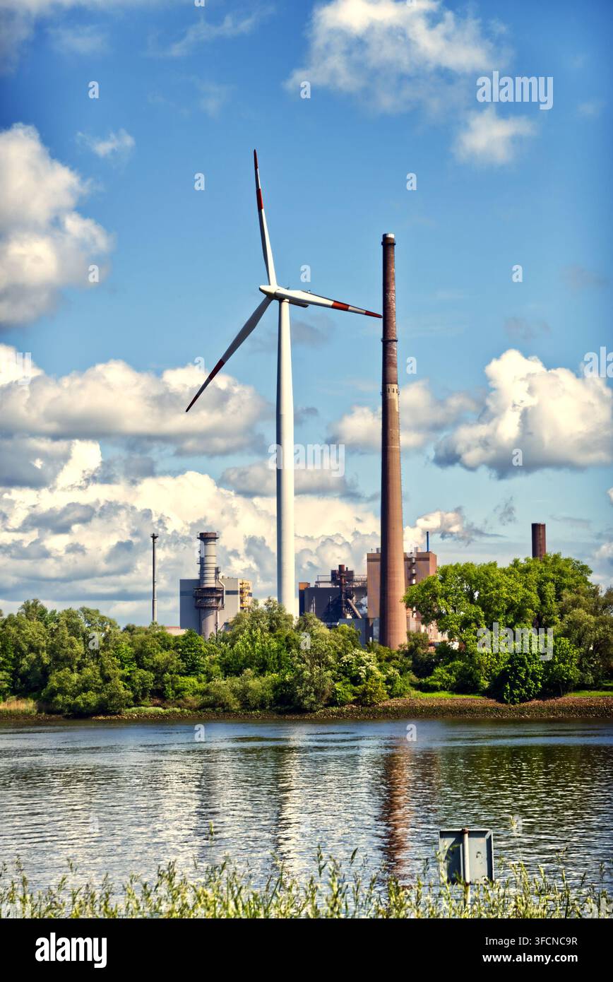 Éolienne et cheminées d'une centrale à combustibles fossiles juxtaposées, représentant l'énergie renouvelable par rapport à l'industrie traditionnelle près d'une rivière Banque D'Images