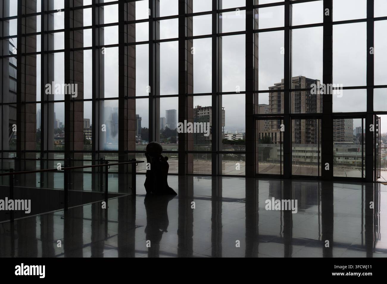 Silhouette d'une personne assise seule dans une pièce moderne aux murs en verre. La skyline de la ville est visible à travers les grandes fenêtres. Le ciel couvert se reflète sur le sol poli. Sc Banque D'Images