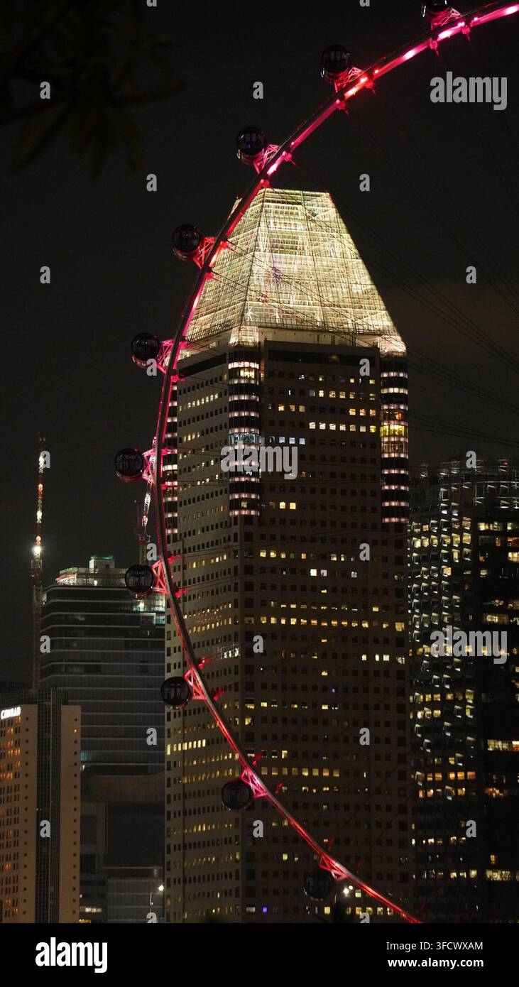Grande roue rouge en arc au-dessus d'une tour pyramidale illuminée la nuit. Un gratte-ciel surmonté d'une pyramide en verre lumineux se dresse à côté des immeubles voisins sous le Banque D'Images