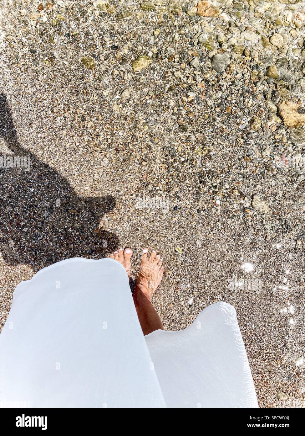 Femme pieds nus en robe blanche debout dans l'eau claire et peu profonde d'une plage de galets - Image de stock capturée avec un smartphone