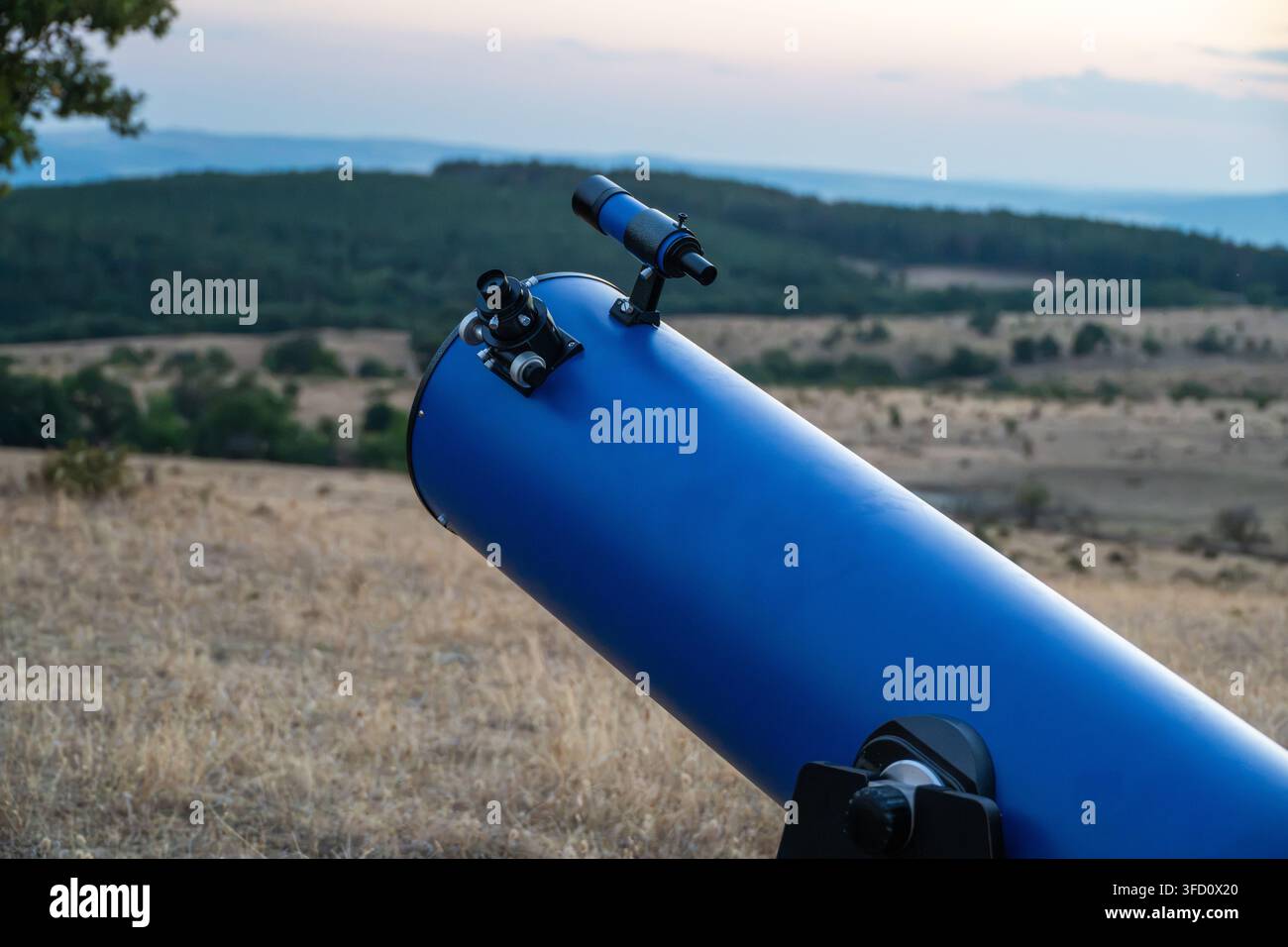 Un télescope à réflexion bleu monté sur une monture Dobson est installé dans un champ sec surplombant un paysage ondulé au coucher du soleil. Banque D'Images