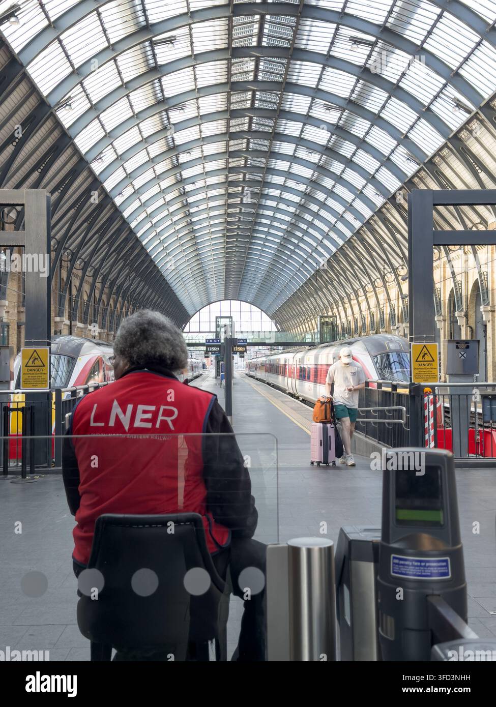Un membre du personnel de la LNER en uniforme rouge aide les passagers à une barrière de billets sur le hall de quai de la gare de Londres King's Cross, Angleterre, Royaume-Uni Banque D'Images