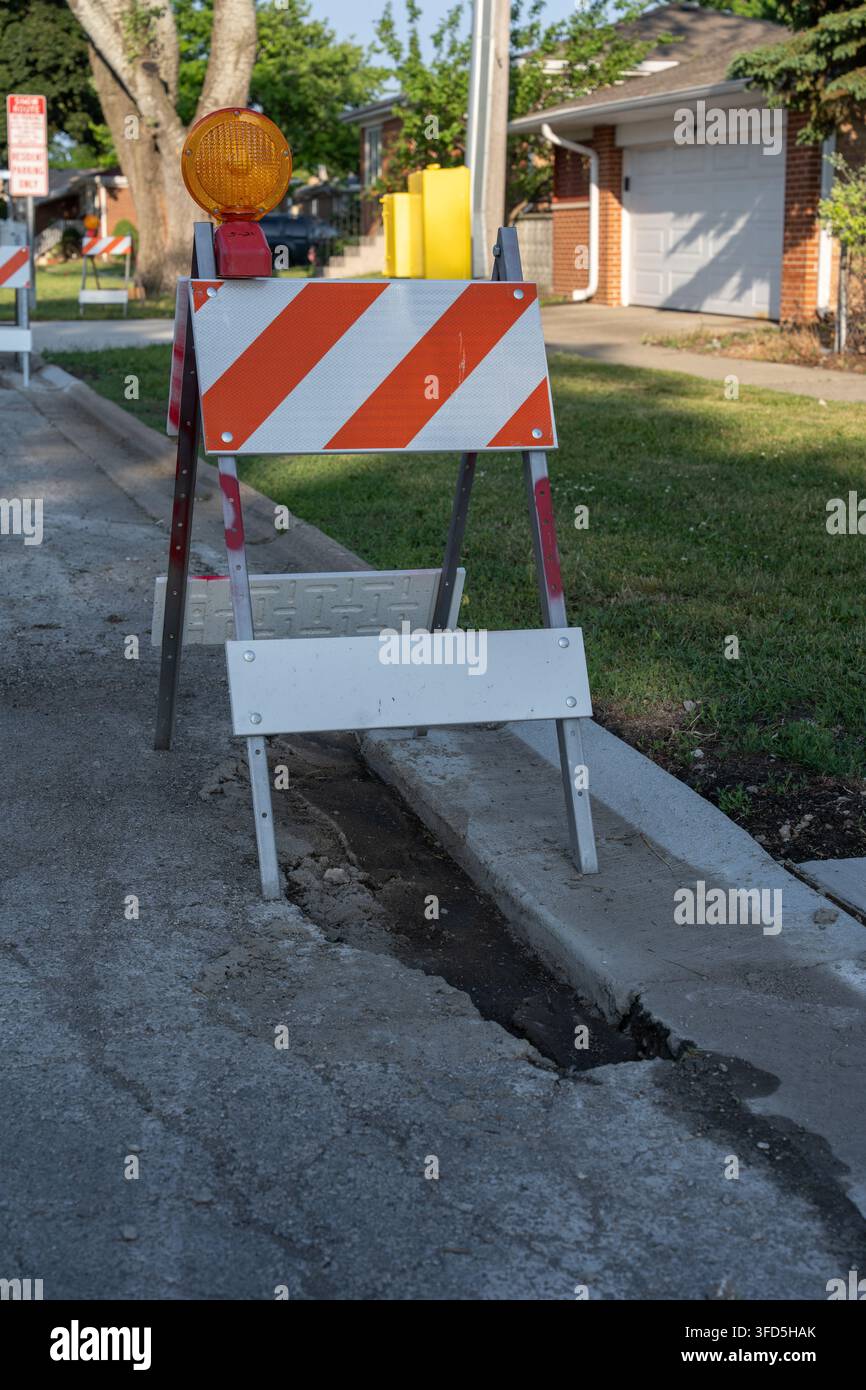 Barrière de circulation orange et blanche avec feu d'avertissement placée à côté d'un trottoir endommagé et d'une zone de travaux sur une rue résidentielle en journée. Banque D'Images