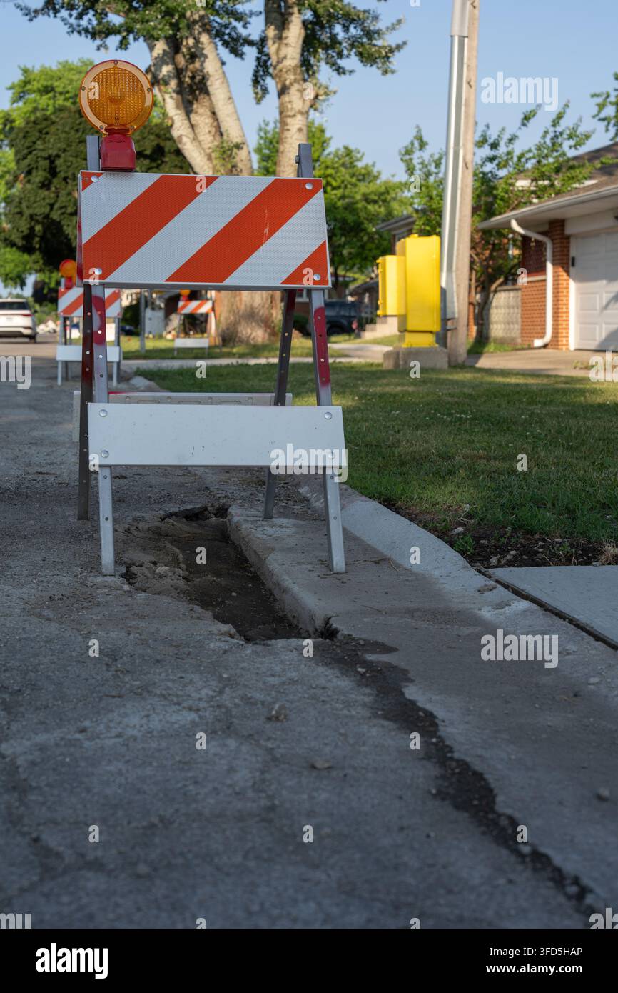 Barrière de circulation orange et blanche avec feu d'avertissement placée à côté d'un trottoir endommagé et d'une zone de travaux sur une rue résidentielle en journée. Banque D'Images