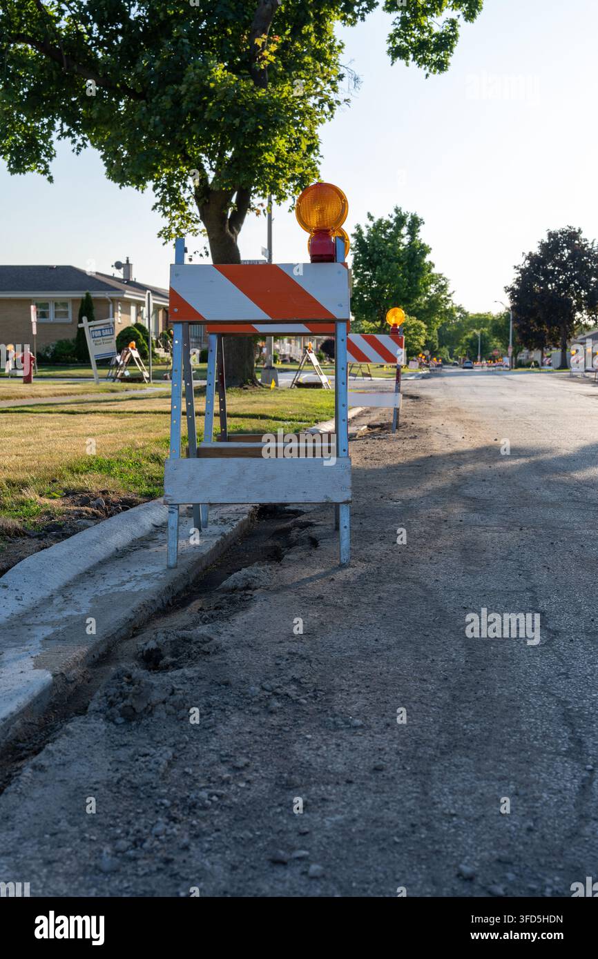 Barrière de circulation orange et blanche avec feu d'avertissement placée à côté d'un trottoir endommagé et d'une zone de travaux sur une rue résidentielle en journée. Banque D'Images