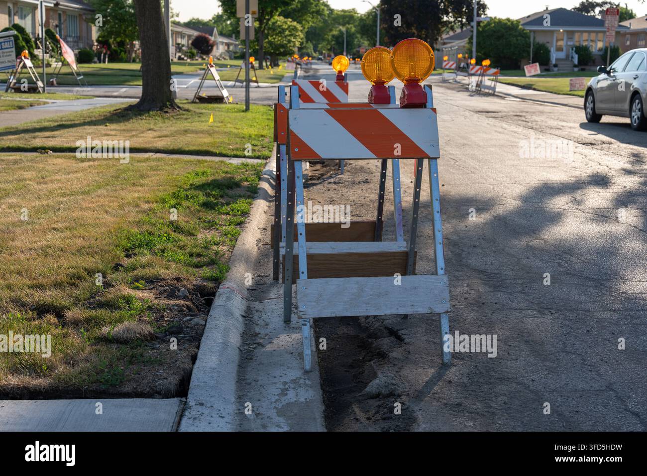 Barrière de circulation orange et blanche avec feu d'avertissement placée à côté d'un trottoir endommagé et d'une zone de travaux sur une rue résidentielle en journée. Banque D'Images