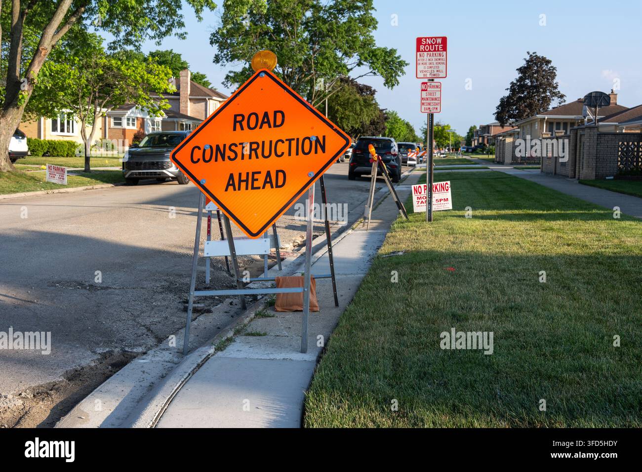 Panneau orange vif indiquant des travaux routiers à venir, placé à côté d'une rue résidentielle avec des barrières de circulation temporaires, des voitures garées et des travaux en cours sur Banque D'Images