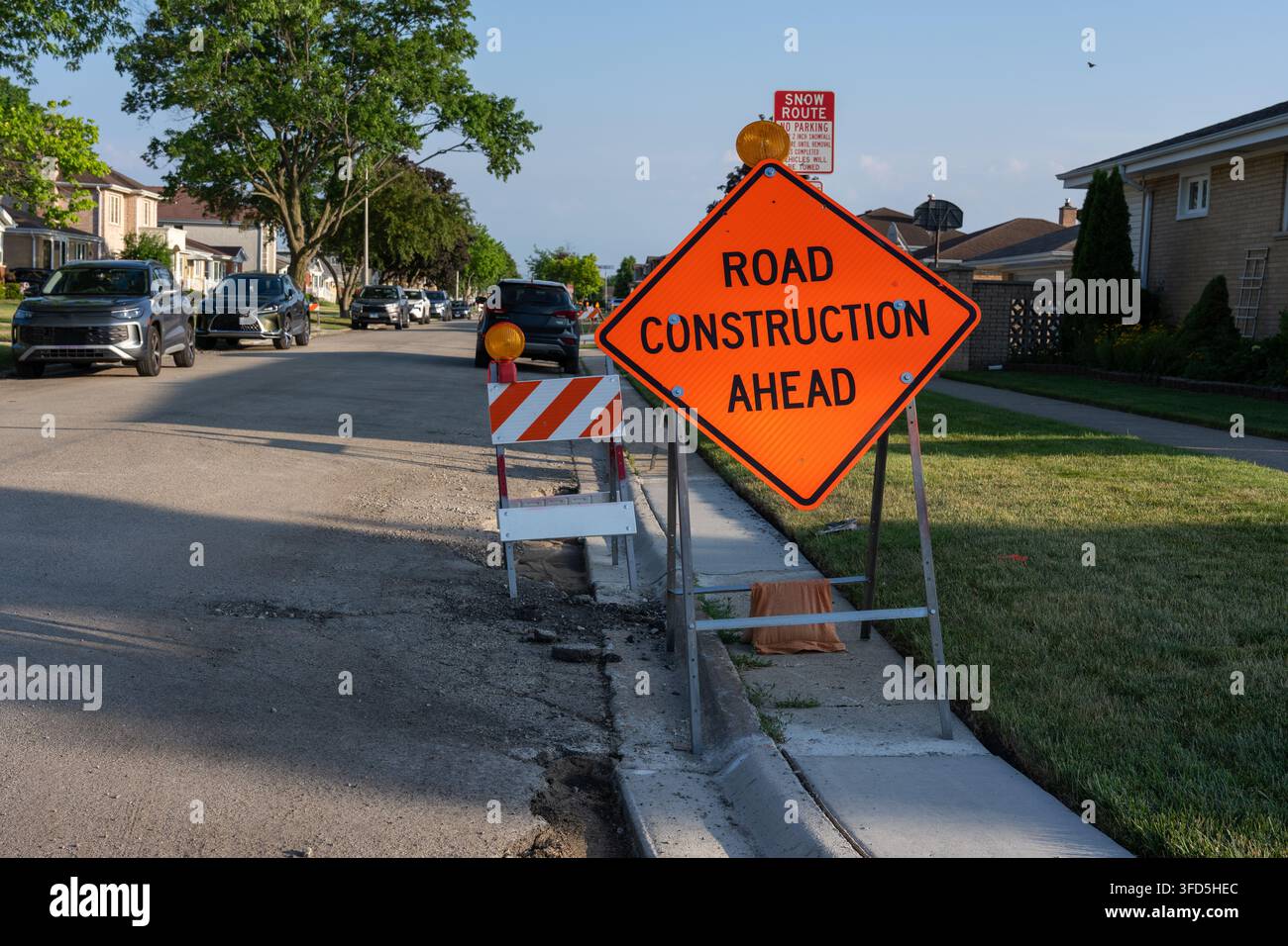 Panneau orange vif indiquant des travaux routiers à venir, placé à côté d'une rue résidentielle avec des barrières de circulation temporaires, des voitures garées et des travaux en cours sur Banque D'Images