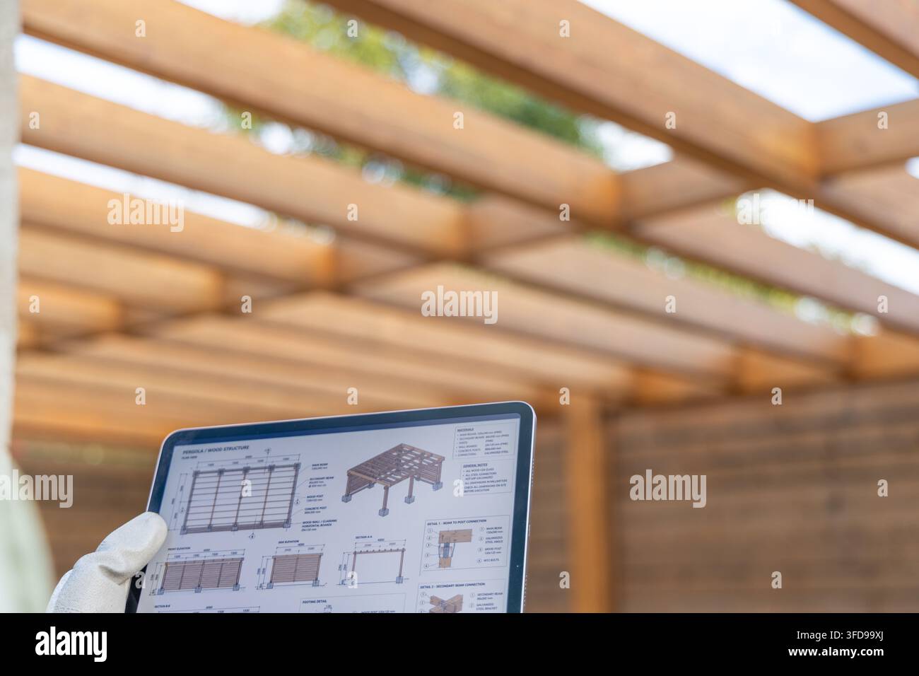 Une main gantée tient une tablette affichant des dessins architecturaux d'une structure en bois, avec un fond flou d'une pergola Banque D'Images