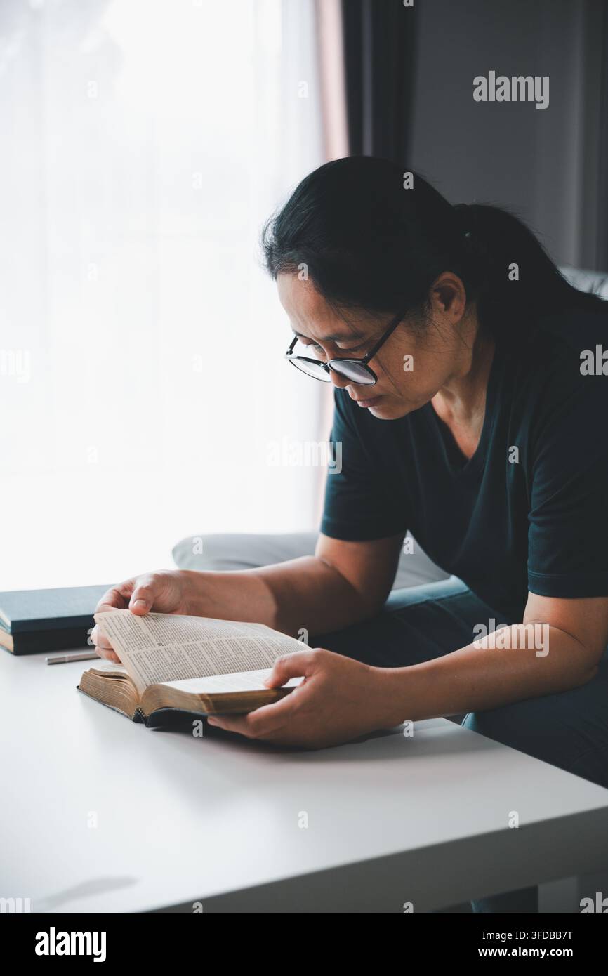 Femme d'âge moyen pensive portant des lunettes et lisant une Bible ouverte à la maison, symbolisant la dévotion chrétienne, l'étude des Écritures, la foi, la sagesse, a Banque D'Images