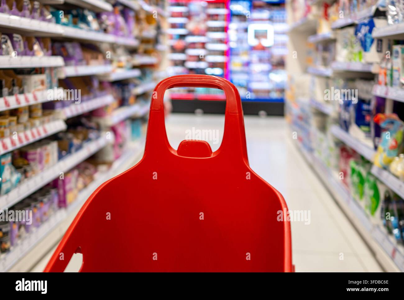 Poignée de chariot rouge avançant dans les allées d'un magasin remplies de produits sur des étagères lumineuses Banque D'Images