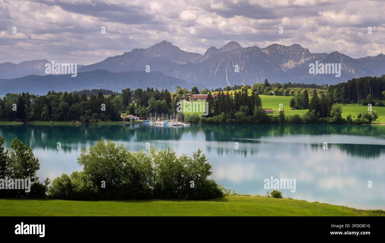 Une vue panoramique du Forggensee révèle une campagne verdoyante, une petite colonie et une chaîne de montagnes alpines escarpées sous des nuages superposés. Arbres et buissons Banque D'Images