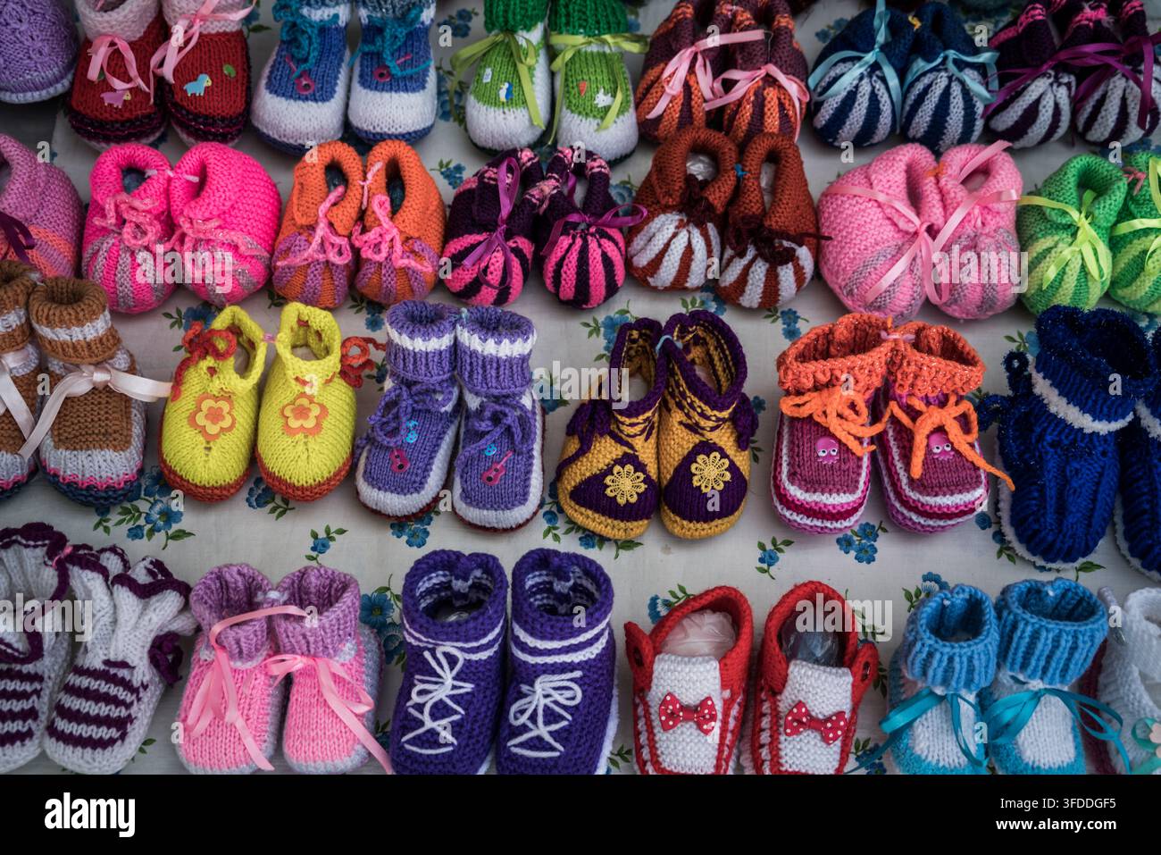 Un éventail vibrant de chaussons et bottines pour bébé tricotés et crochetés à la main est soigneusement disposé sur une surface en tissu blanc avec un motif floral bleu. Banque D'Images