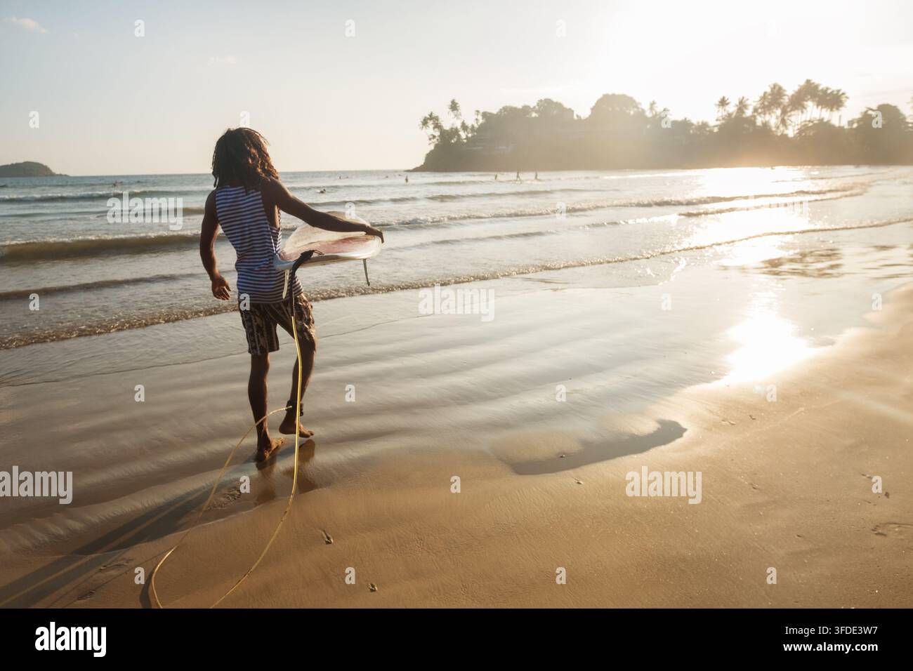 Homme noir aux cheveux longs tenant une planche de surf blanche, marchant sur le sable mouillé vers les vagues de l'océan à la belle plage de Dewata au Sri Lanka pendant la chaude heure dorée Banque D'Images
