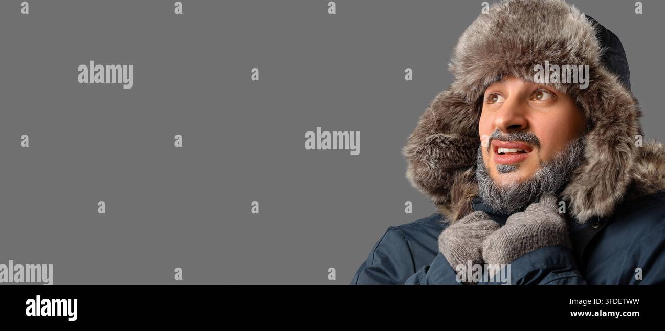 Froid extrême et survie hivernale, homme portant un chapeau en fourrure et un parkas frissonnant à des températures glaciales avec du givre sur la barbe devant un fond gris Banque D'Images