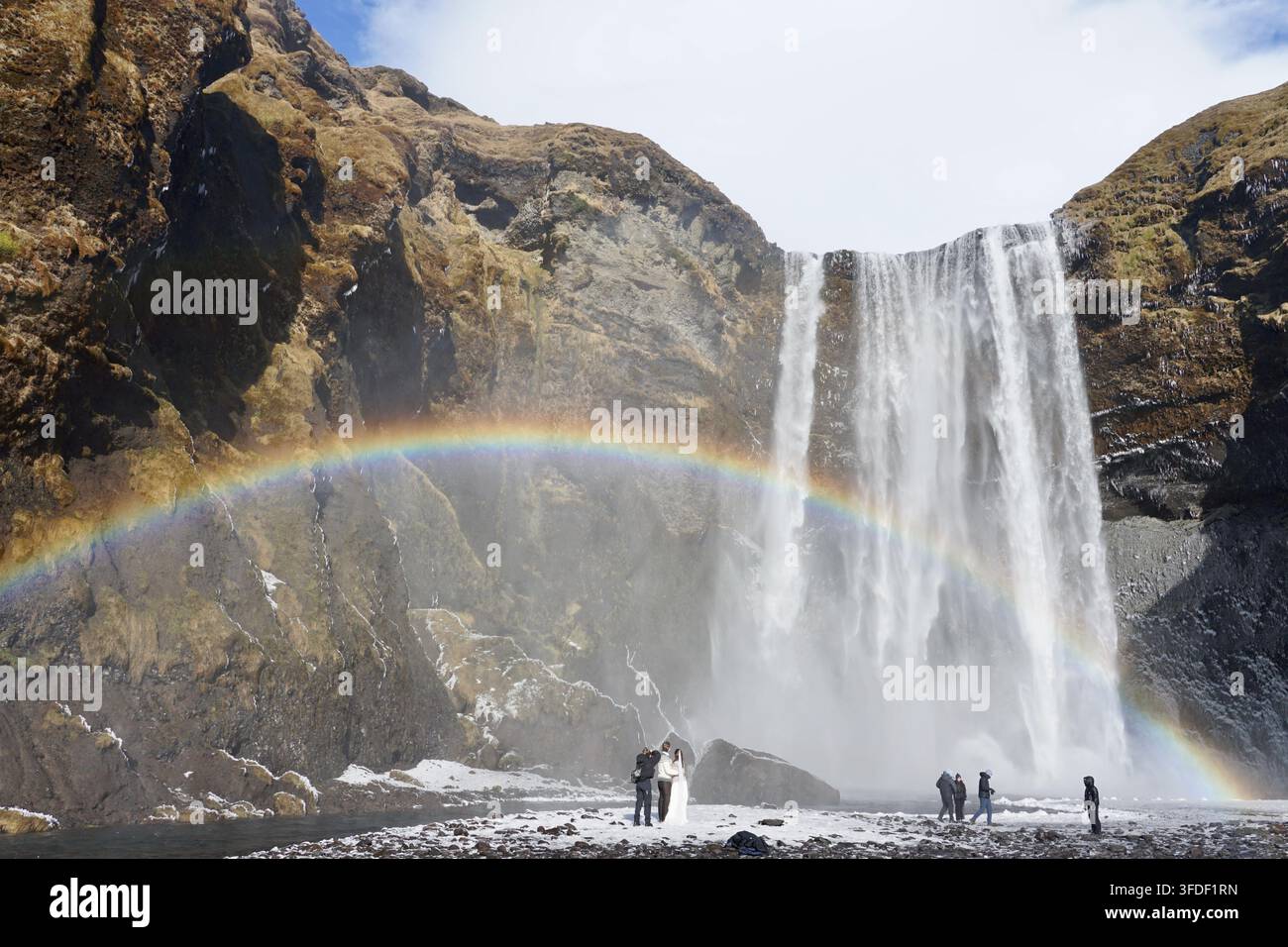 Une mariée et un marié posent pour des photos près d'une puissante cascade avec un arc-en-ciel éclatant enjambant des falaises brumeuses, tandis que les photographes et les spectateurs capturent le paysage côtier dramatique islandais. Banque D'Images