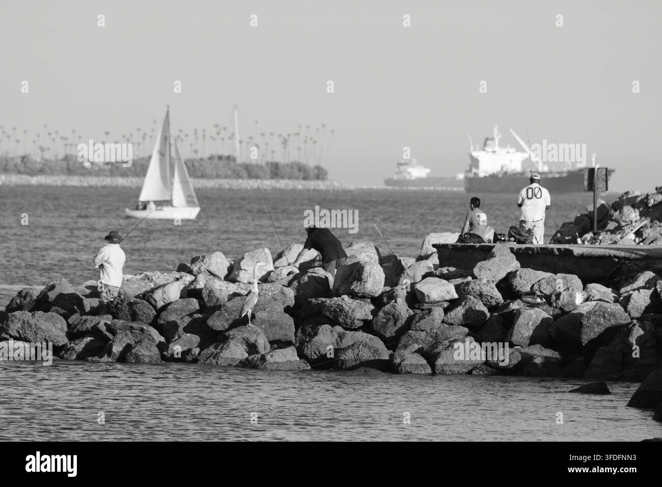 Les pêcheurs pêchent depuis les rochers tandis qu'un voilier et des cargos traversent le port. Banque D'Images