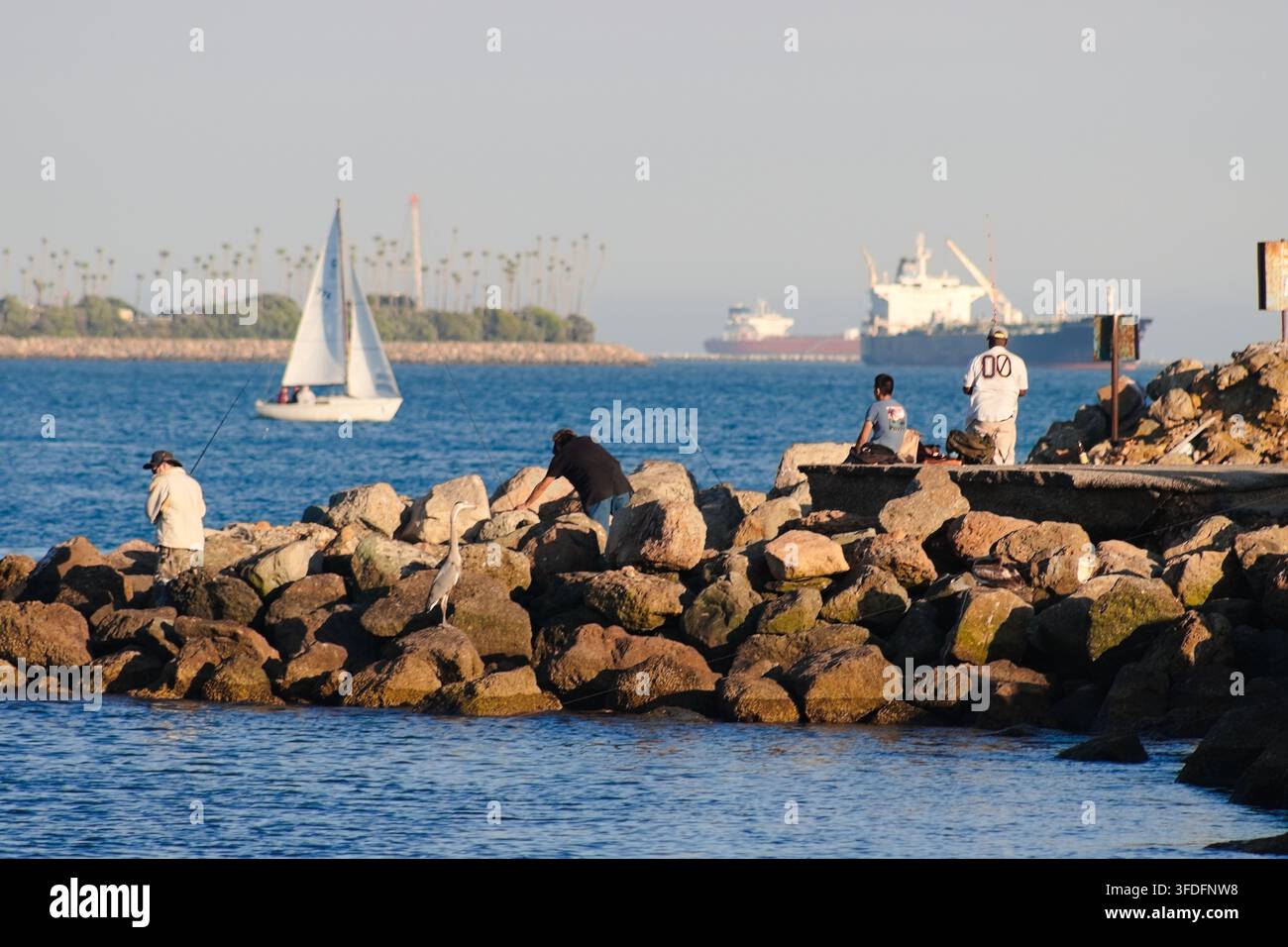 Les pêcheurs pêchent depuis les rochers tandis qu'un voilier et des cargos traversent le port. Banque D'Images