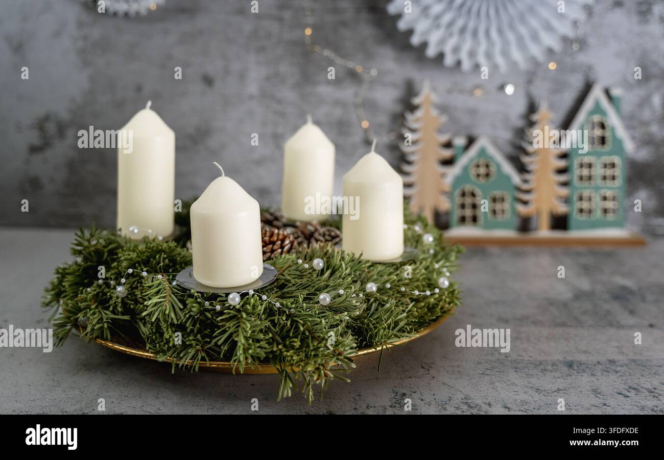 Couronne de l'Avent traditionnelle disposée avec quatre bougies blanches et des branches de feuillus luxuriantes devant un village de Noël miniature créant une ambiance hivernale chaleureuse Banque D'Images