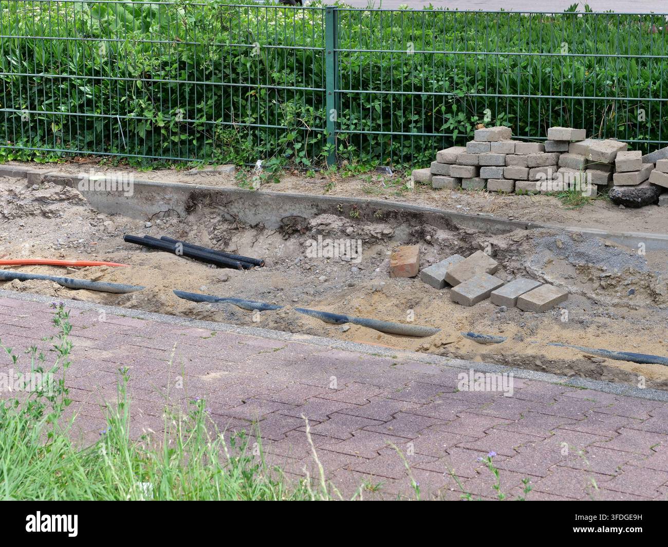 Vue rapprochée d'un chantier de construction de rue montrant une tranchée creusée avec un câblage électrique exposé et des lignes en fibre optique orange. Les pavés sont s Banque D'Images
