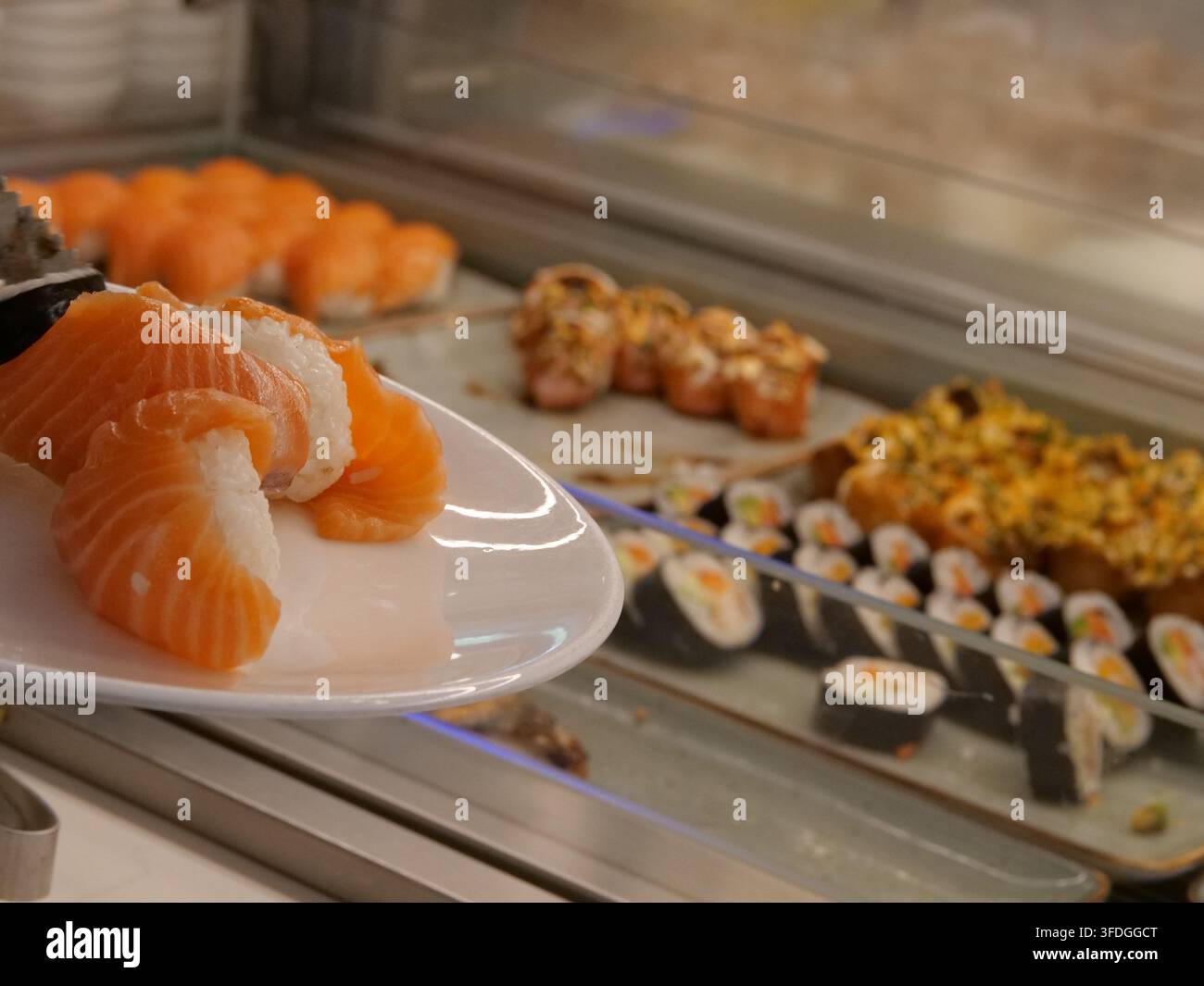 Prise de vue concentrée sur un délicieux sushi nigiri au saumon servi dans une assiette en porcelaine blanche tenue par un client. Le fond flou montre une grande variété de fré Banque D'Images